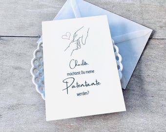 May include: A white card with a black line drawing of two hands holding each other. The text on the card reads "Charlotte, möchtest Du meine Patenante werden?" which translates to "Charlotte, would you like to be my godmother?"