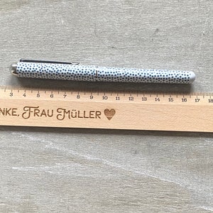 K&ouml;nnte beinhalten: Ein Holzlineal mit einem Herz und dem eingravierten Text "DANKE, FRAU M&Uuml;LLER". Ein wei&szlig;-schwarzer Stift mit Punkten liegt auf dem Lineal.