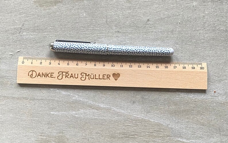 K&ouml;nnte beinhalten: Ein Holzlineal mit einem Herz und dem eingravierten Text "DANKE, FRAU M&Uuml;LLER". Ein wei&szlig;-schwarzer Stift mit Punkten liegt auf dem Lineal.