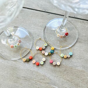 May include: Set of eight colorful glass charms with names spelled out in gold letters. The charms are designed to hang from wine glasses.