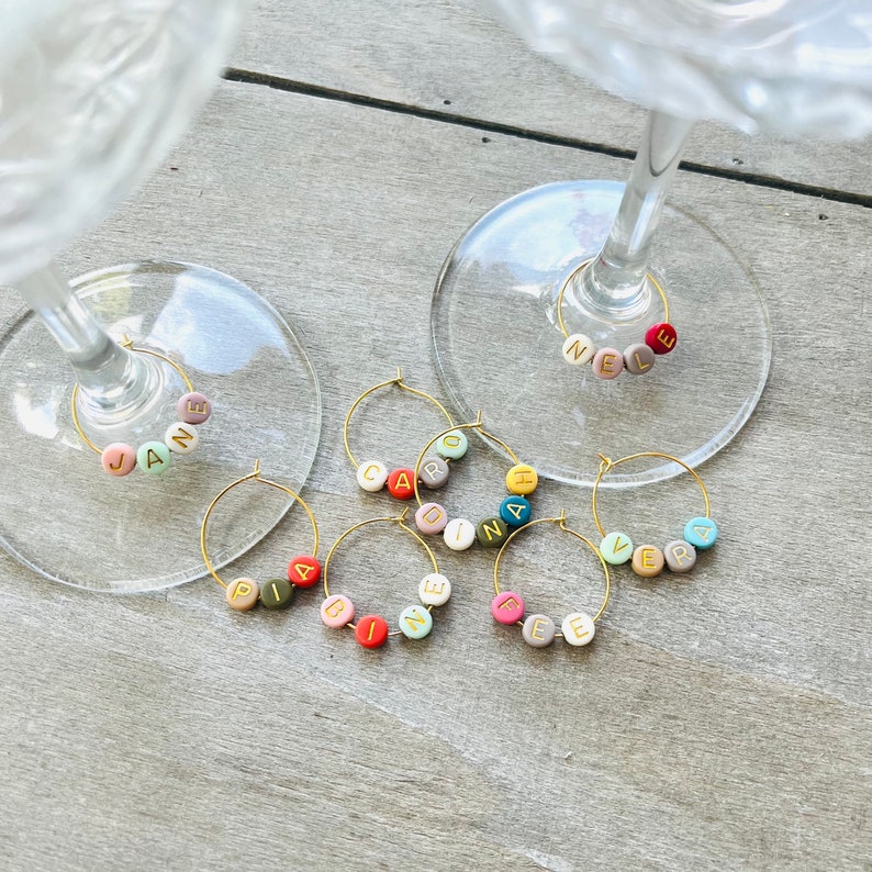 May include: Set of eight colorful glass charms with names spelled out in gold letters. The charms are designed to hang from wine glasses.