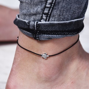 Könnte beinhalten: Ein schwarzes Fußkettchen mit einem kleinen silbernen Schildkrötenanhänger.