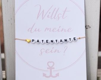 May include: A greeting card with the text "Willst du meine PATENTANTE sein?" in pink script, with a bracelet featuring letter beads spelling "PATENTANTE" and a gold heart charm. The card has a pink anchor illustration.