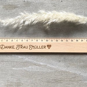 K&ouml;nnte beinhalten: Ein Holzlineal mit einem schwarzen Herz und dem Text "DANKE, FRAU M&Uuml;LLER" eingraviert. Das Lineal ist 50,8 cm lang und hat Markierungen f&uuml;r jeden Zentimeter.
