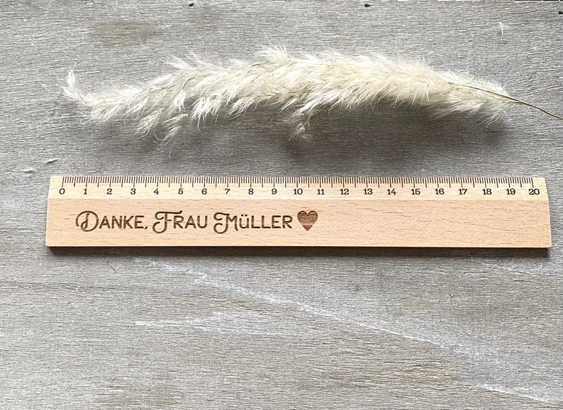 K&ouml;nnte beinhalten: Ein Holzlineal mit einem schwarzen Herz und dem Text "DANKE, FRAU M&Uuml;LLER" eingraviert. Das Lineal ist 50,8 cm lang und hat Markierungen f&uuml;r jeden Zentimeter.