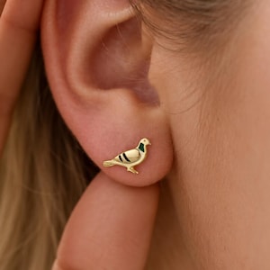 May include: A small gold stud earring shaped like a pigeon. The bird figure features a polished gold finish with a distinct green accent on the neck and two black horizontal stripes on the wing, securely positioned on the earlobe for display.