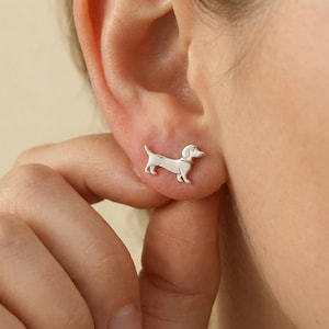 Könnte beinhalten: Nahaufnahme eines silbernen Dackel-Ohrsteckers. Der Ohrring ist eine detaillierte Miniaturdarstellung eines Dackels mit Halsband und leicht erhobenem Kopf. Der Ohrring wird an einem Ohr getragen.