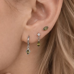 May include: A set of three silver-toned earrings featuring marquise-cut olive green stones. One earring is a small hoop with a dangling stone, another is a stud with a dangling stone and a clear accent, and the third is a simple marquise stud.
