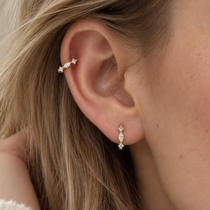 Op de afbeelding: Een gouden oorklem en een bijpassende gouden oorring, beide met een verfijnd ontwerp met kleine, heldere, marquise- en rondgeslepen stenen in een verticale rij, gedragen op het bovenste kraakbeen en in de oorlel.