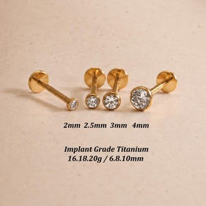 May include: Four gold-colored labret studs with clear round stones in bezel settings, arranged by size. Text below reads: 2mm 2.5mm 3mm 4mm, Implant Grade Titanium, 16.18.20g / 6.8.10mm. The studs feature flat back posts and circular front gems.