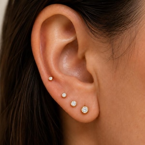May include: A set of four gold stud earrings with clear round stones, arranged in a line along the earlobe and cartilage. Each earring features a bezel setting that holds a single sparkling stone, graduating in size from smallest to largest.