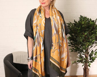 May include: A woman wearing a gray linen tunic with a yellow and green scarf featuring a bird print. The scarf has a green tassel and a blue and yellow stripe. She is wearing white pants and nude sandals.