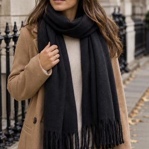 May include: A long black scarf with a thick knit texture and fringed ends, worn over a beige sweater and a tan wool coat. The scarf is wrapped around the neck, featuring a soft, dense fabric construction with visible vertical weave patterns.
