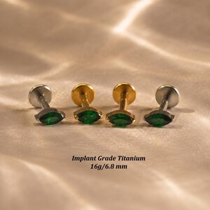 May include: Four labret studs featuring marquise-cut green gemstones. Two studs have a silver-tone finish and two have a gold-tone finish. The text Implant Grade Titanium 16g/6.8 mm is visible below the jewelry, indicating the material and size.