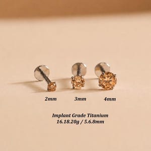 May include: Three implant grade titanium labret studs with round champagne-colored gemstones in 2mm, 3mm, and 4mm sizes. The text reads: 2mm, 3mm, 4mm, Implant Grade Titanium, 16.18.20g / 5.6.8mm. Each stud features a flat back and prong setting.