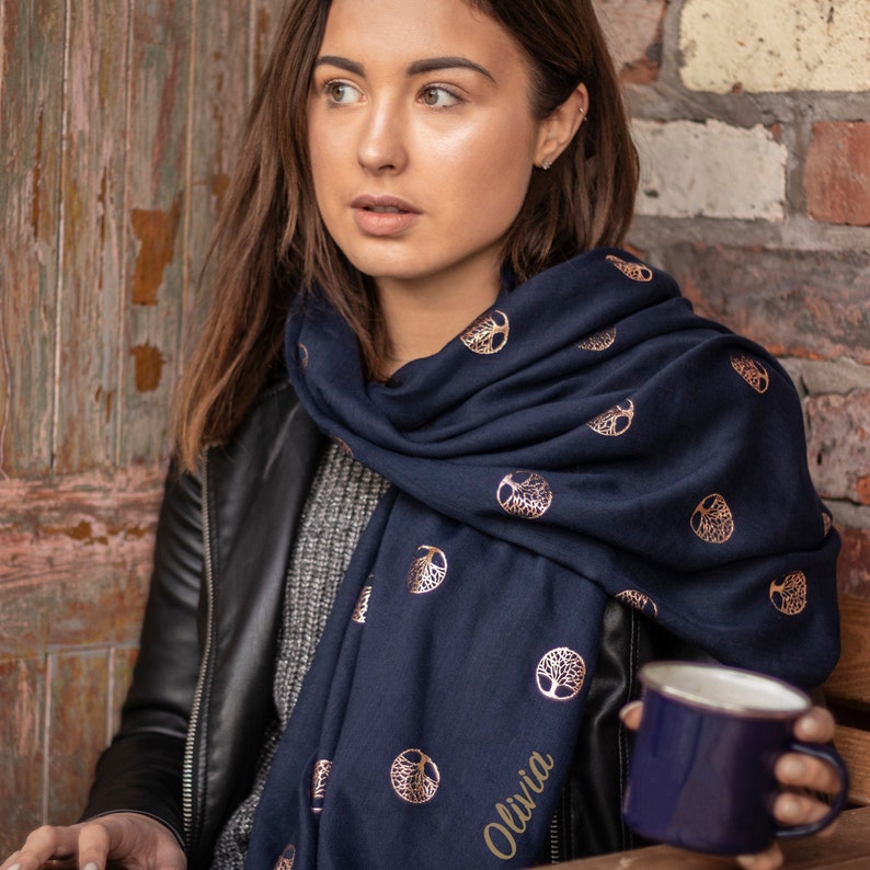model wearing a navy blue lightweight scarf around the neck, the scarf has an all over gold foil tree of life pattern, the trees are encased in a circle.