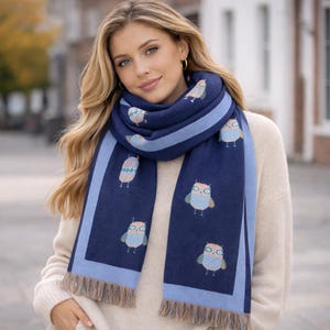 Puede incluir: Bufanda azul marino con borde y flecos azul claro. La bufanda presenta un patrón repetido de ilustraciones de búhos coloridos. La mujer lleva un jersey color crema.