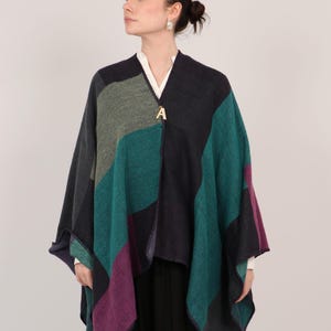 May include: A patchwork poncho in shades of teal, purple, gray, and navy blue. The poncho has a gold-colored letter "A" pendant at the neckline. The garment is worn over a white shirt and black skirt.