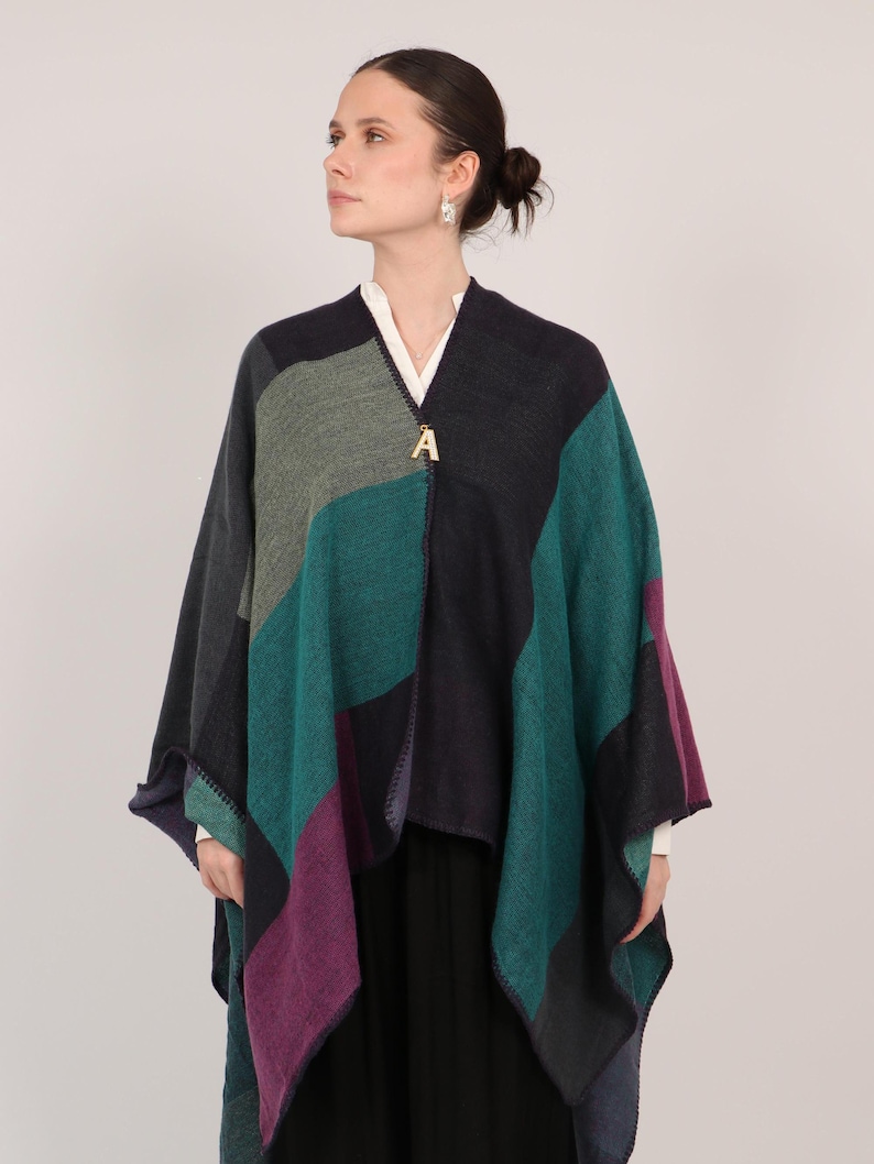 May include: A patchwork poncho in shades of teal, purple, gray, and navy blue. The poncho has a gold-colored letter "A" pendant at the neckline. The garment is worn over a white shirt and black skirt.