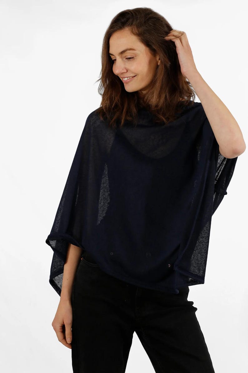 May include: Navy blue poncho-style top with a loose, flowing design. The top has a cowl neck and is made of a sheer, lightweight fabric. The garment is worn over a black top and paired with black pants.
