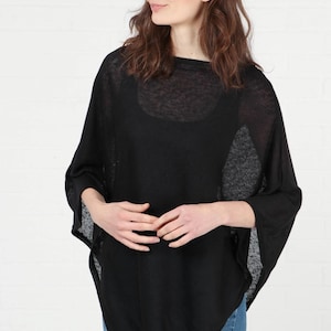 May include: A black poncho with a round neckline and three-quarter sleeves. The lightweight fabric drapes loosely, creating a relaxed silhouette. The poncho is worn over a black top and paired with blue jeans.