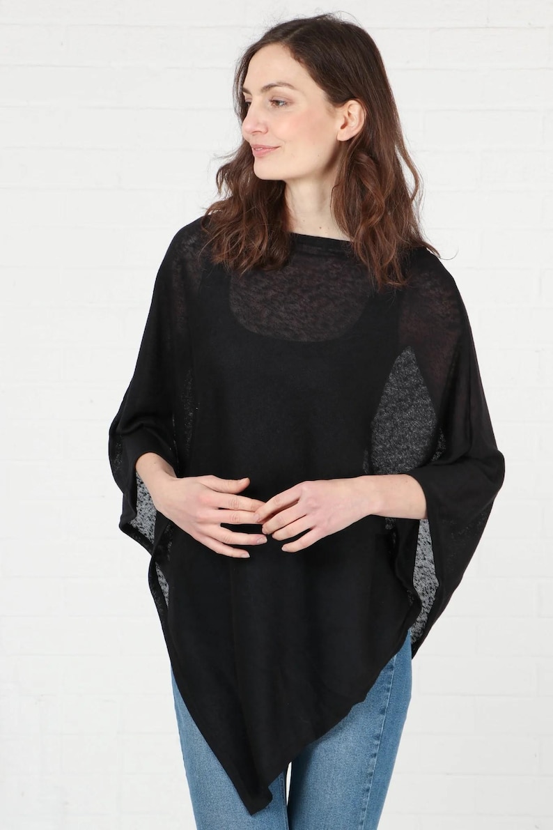 May include: A black poncho with a round neckline and three-quarter sleeves. The lightweight fabric drapes loosely, creating a relaxed silhouette. The poncho is worn over a black top and paired with blue jeans.