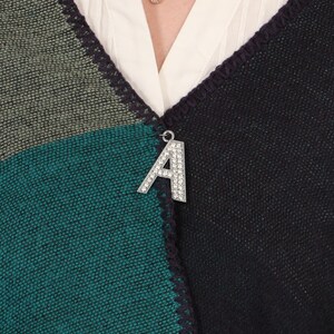 May include: A color-blocked shawl in shades of teal, olive green, and black, with a decorative silver letter "A" brooch. The shawl has a textured appearance and is worn over a white blouse.