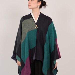 May include: A multi-colored knitted poncho with a geometric pattern in shades of teal, purple, gray, and black. The poncho has a gold letter "A" charm attached to the front.