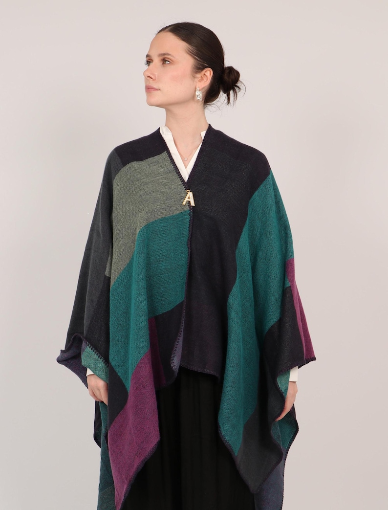 May include: A multi-colored knitted poncho with a geometric pattern in shades of teal, purple, gray, and black. The poncho has a gold letter "A" charm attached to the front.