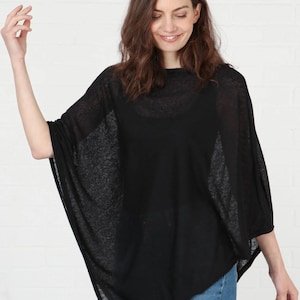 May include: A black poncho-style top with a loose, flowing design. The top is made of a sheer fabric and is worn with blue jeans. The model is smiling and standing in front of a white brick wall.