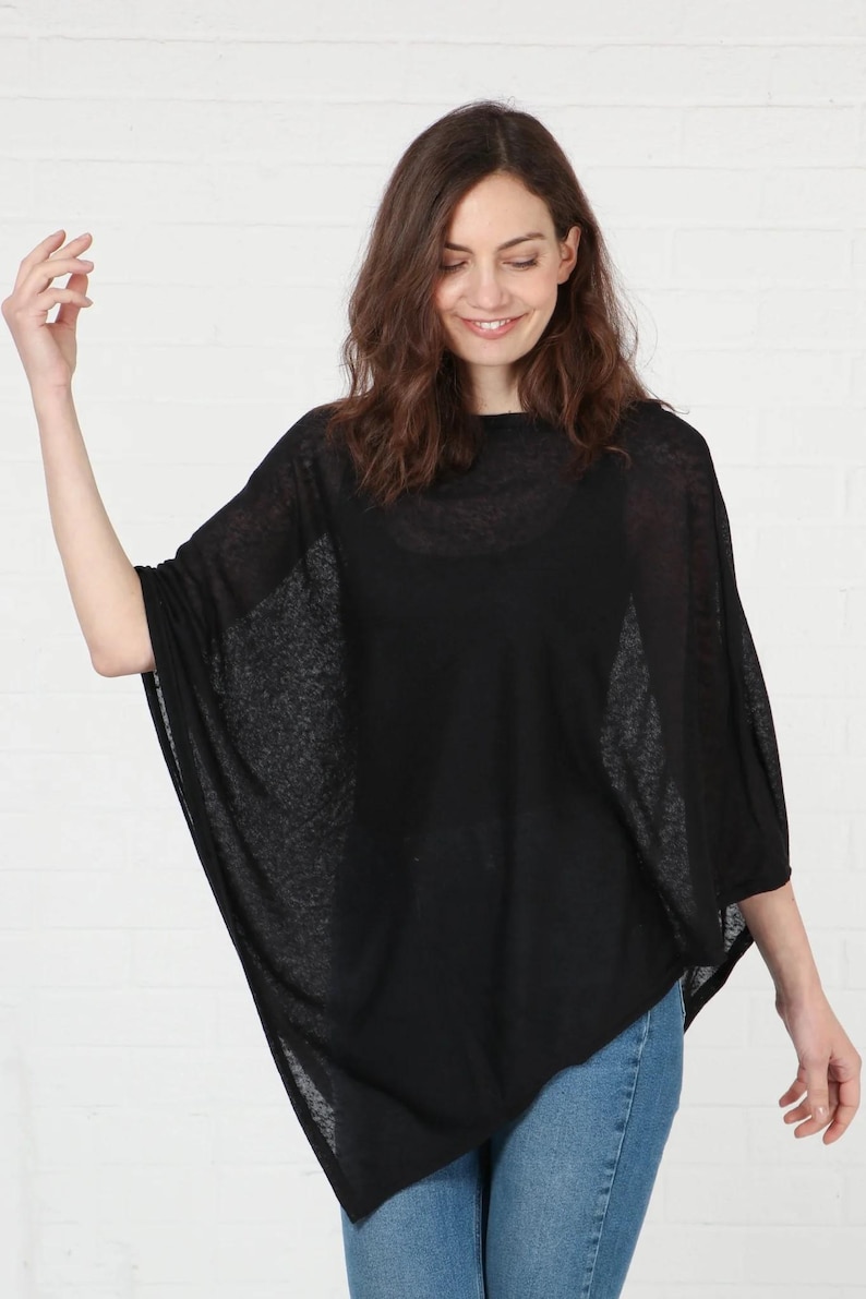 May include: A black poncho-style top with a loose, flowing design. The top is made of a sheer fabric and is worn with blue jeans. The model is smiling and standing in front of a white brick wall.
