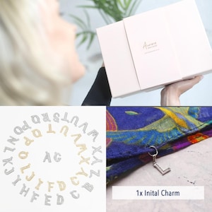 May include: A close-up of a silver initial charm in the shape of the letter L, hanging from a blue and yellow patterned scarf. The charm is part of a set of 26 initial charms, shown in a circle around the scarf. The charms are available in silver and gold.