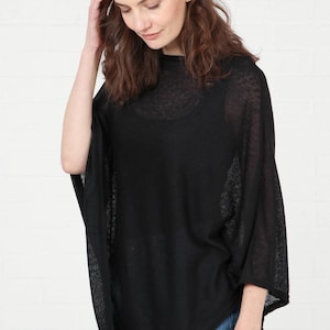 May include: Black poncho-style top with a semi-sheer, lightweight fabric. The top has a relaxed fit with wide sleeves and a high neckline. The model is wearing blue jeans. This versatile garment can be worn for various occasions.