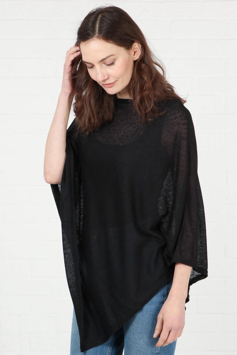 May include: Black poncho-style top with a semi-sheer, lightweight fabric. The top has a relaxed fit with wide sleeves and a high neckline. The model is wearing blue jeans. This versatile garment can be worn for various occasions.