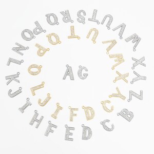 May include: A collection of silver and gold-toned alphabet letter charms, each encrusted with small, sparkling crystals. The letters are arranged in a circular pattern, with two letters, "AG", in the center. These charms are ideal for jewelry making.