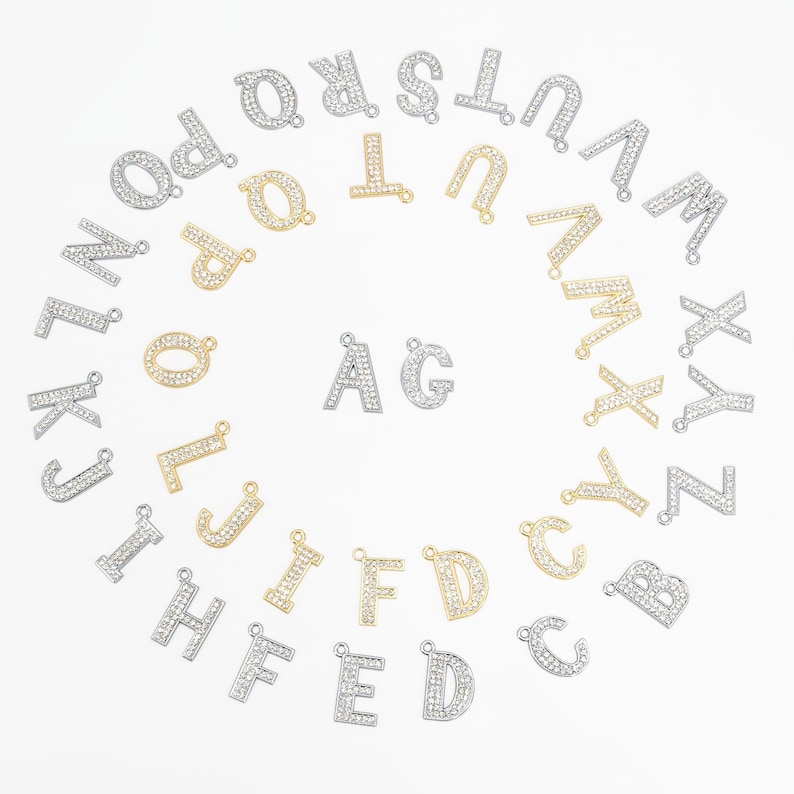 May include: A collection of silver and gold-toned alphabet letter charms, each encrusted with small, sparkling crystals. The letters are arranged in a circular pattern, with two letters, "AG", in the center. These charms are ideal for jewelry making.