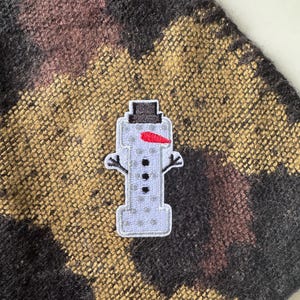 May include: A snowman patch with a black top hat, a red carrot nose, and three black buttons. The patch is on a textured fabric with a pattern of black, brown, and gold.