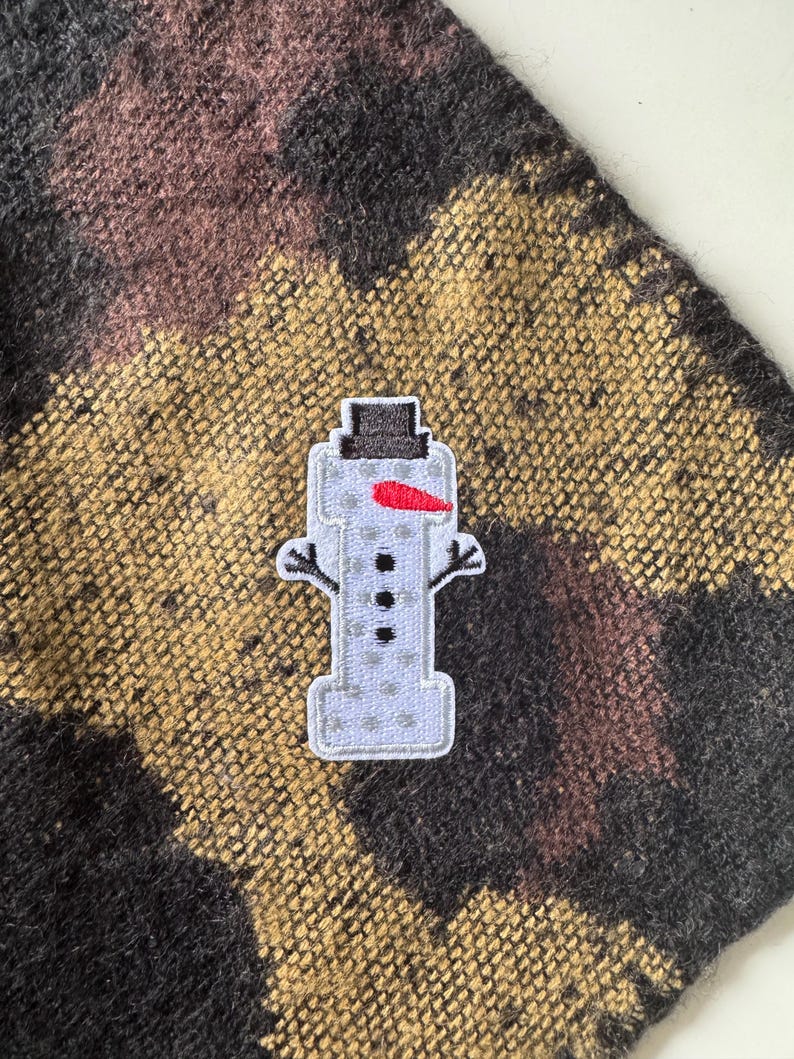 May include: A snowman patch with a black top hat, a red carrot nose, and three black buttons. The patch is on a textured fabric with a pattern of black, brown, and gold.