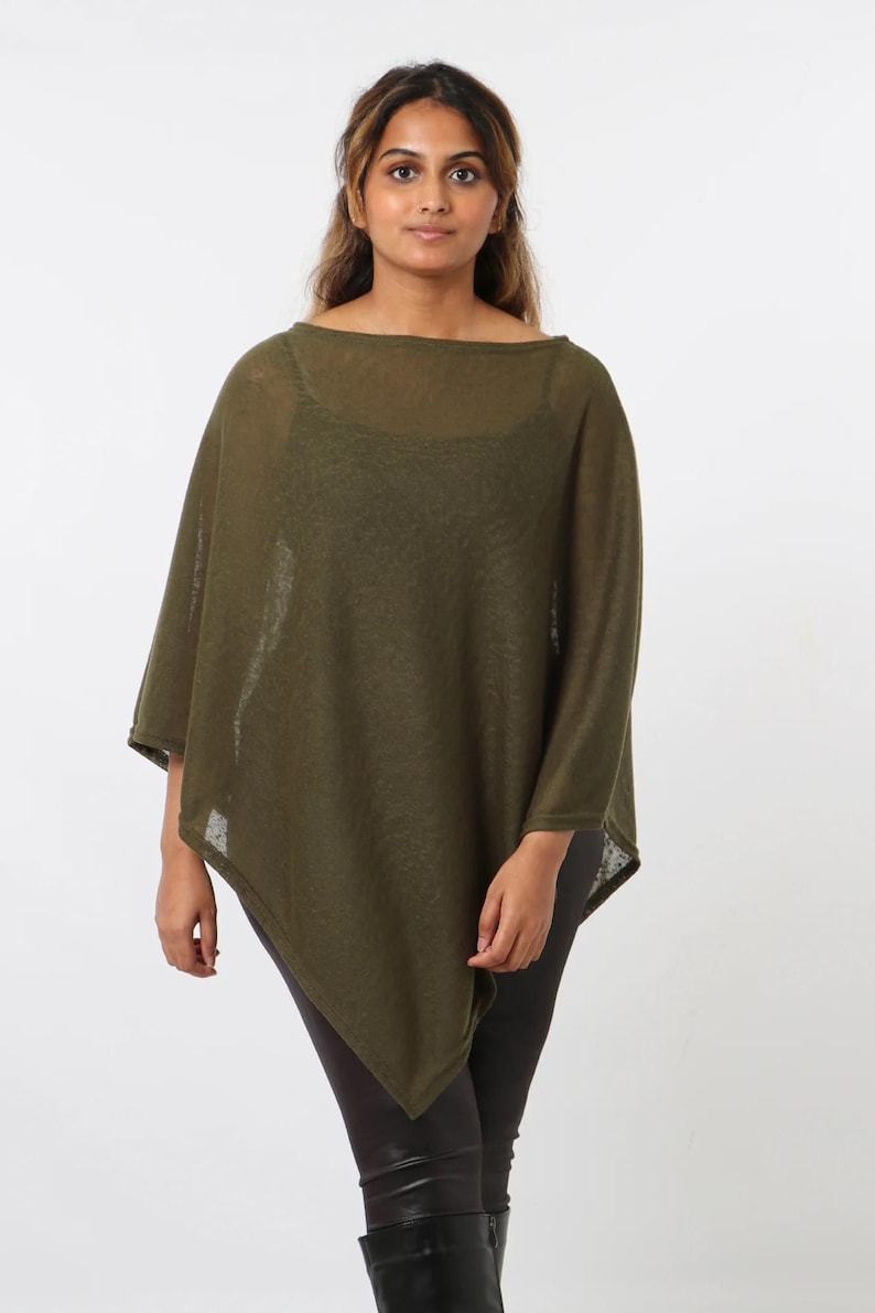 May include: An olive green poncho with a boat neck and asymmetrical hemline. The lightweight knitwear garment is worn over a black top and dark leggings. The poncho is a versatile piece of clothing.