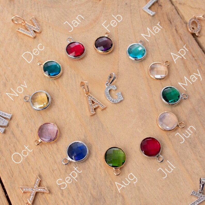 Pu&ograve; includere: Una collezione di 12 ciondoli in oro e argento con pietre preziose colorate. I ciondoli hanno la forma di lettere, mesi dell'anno e altri disegni. I ciondoli sono disposti su una superficie di legno.