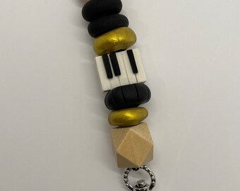 May include: A lanyard with a musical theme. The lanyard features a series of beads in various shapes and colors, including black, gold, and a piano key design. A silver-colored clasp is attached at the bottom, and a black cord at the top.