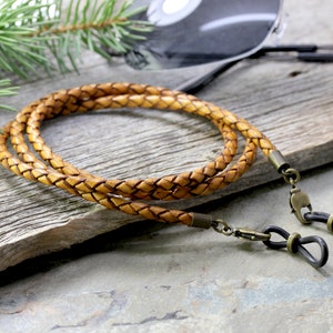 May include: A braided leather eyeglass strap with gold-tone metal clasps and black rubber tips. The strap is light brown and has a woven texture.