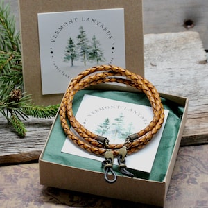 May include: A braided leather eyeglass lanyard in a gift box. The light brown lanyard has bronze-colored clasps and is designed to hold eyeglasses. The box contains a card with the text "Vermont Lanyards" and a watercolor illustration of pine trees.