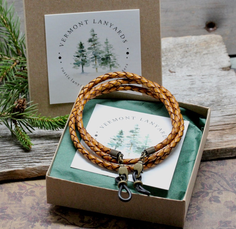 May include: A braided leather eyeglass lanyard in a gift box. The light brown lanyard has bronze-colored clasps and is designed to hold eyeglasses. The box contains a card with the text "Vermont Lanyards" and a watercolor illustration of pine trees.
