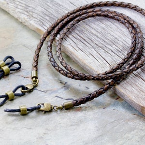 May include: Braided brown leather eyeglass chain with brass-colored clasps and loops. The chain is designed to hold eyeglasses and features a woven texture. Includes a pair of black rubber loops.