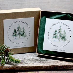 May include: Two kraft paper boxes, one open, with a white label featuring a watercolor image of pine trees. The text "VERMONT LANYARDS" is above the trees, and "rustic lanyards & eyeglass holders" is below. The website is also listed.