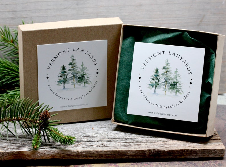 May include: Two kraft paper boxes, one open, with a white label featuring a watercolor image of pine trees. The text "VERMONT LANYARDS" is above the trees, and "rustic lanyards & eyeglass holders" is below. The website is also listed.