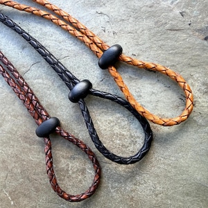 May include: Three braided leather cords in tan, black, and dark brown. Each cord features a smooth, matte black oval bead that acts as a sliding toggle to adjust the size of the loop at the end of the braided leather strand.