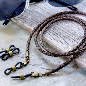 May include: Braided brown leather eyeglass chain with brass-colored clasps and connectors. The chain is next to a pair of black sunglasses and a set of black eyeglass holders with brass accents. The chain is approximately 30 inches long.