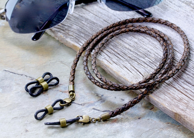 May include: Braided brown leather eyeglass chain with brass-colored clasps and connectors. The chain is next to a pair of black sunglasses and a set of black eyeglass holders with brass accents. The chain is approximately 30 inches long.